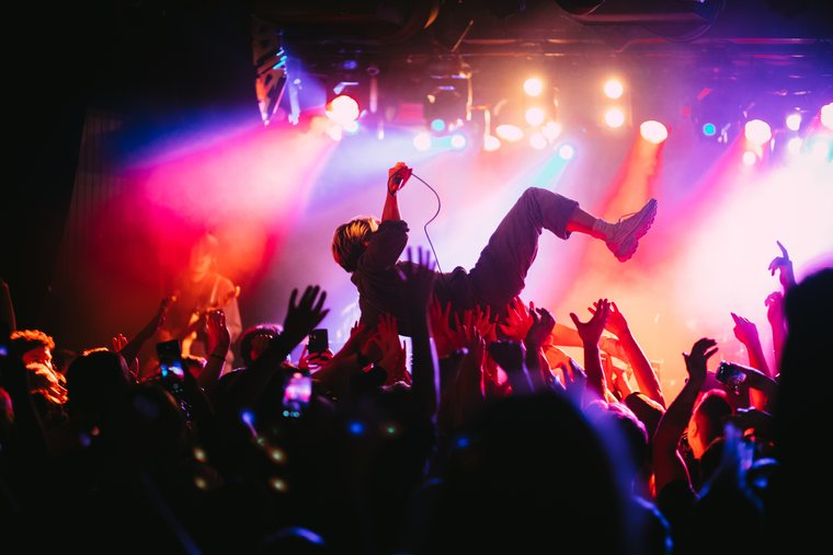 A performer crowd-surfs above raised hands in vivid pink and blue light.