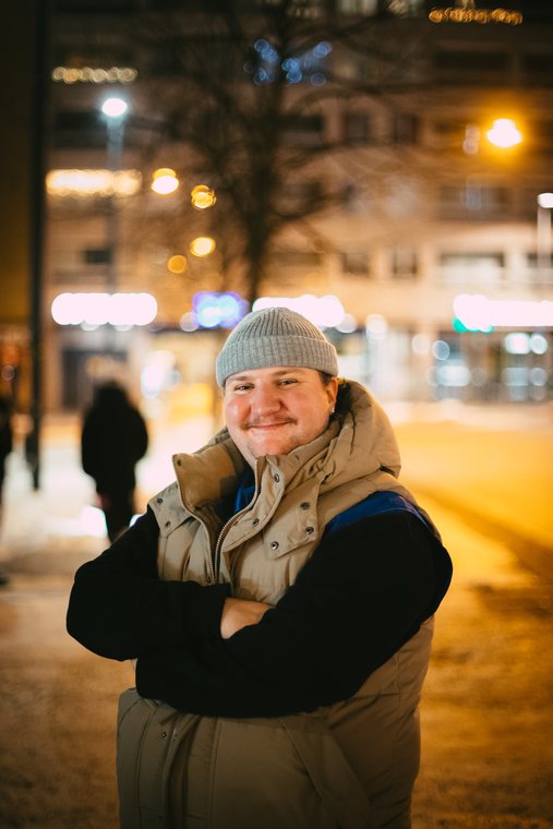 A smiling person in a knit cap poses on a softly lit winter street.