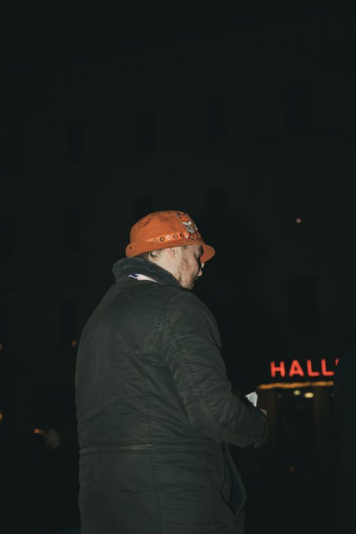 A person in a cap stands at night beside a glowing red hall sign.