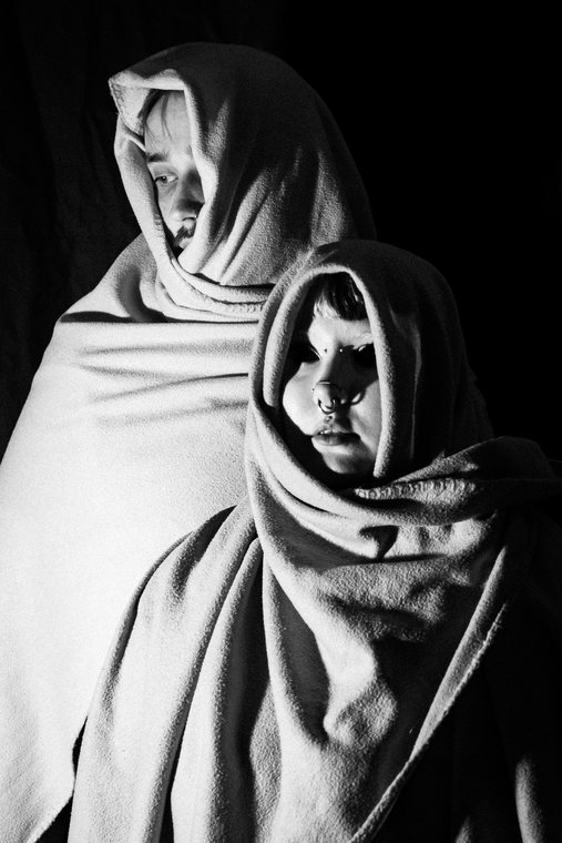 Two figures wrapped in pale fabric stand together against a black background.