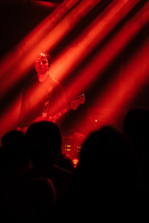 A performer stands beneath diagonal red beams before a dark audience.